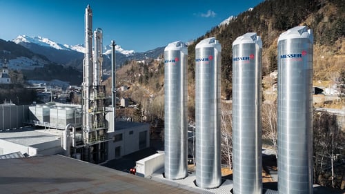 Cuatro altos depósitos industriales de gas con la palabra «Messer» se alzan junto a una planta de captura de carbono en una zona montañosa, bajo un cielo azul brillante.